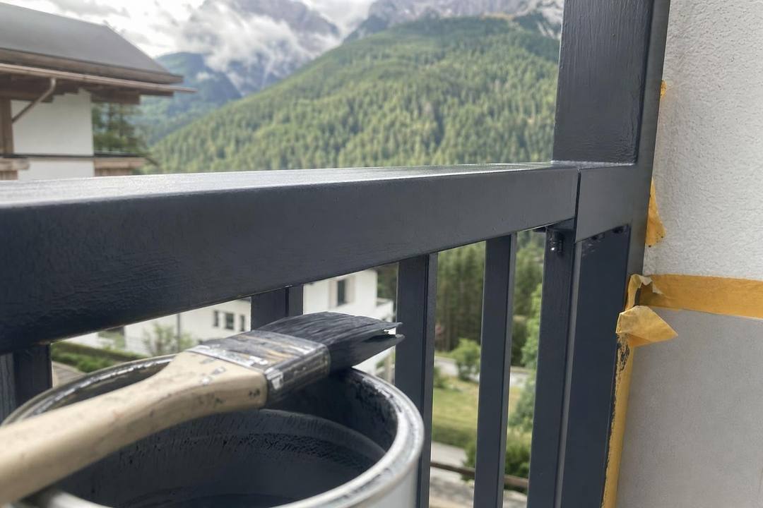Balkongeländer aus Metall in dunklem Anthrazit, frisch gestrichen; daneben Pinsel, Farbtopf und abgeklebte Wand.