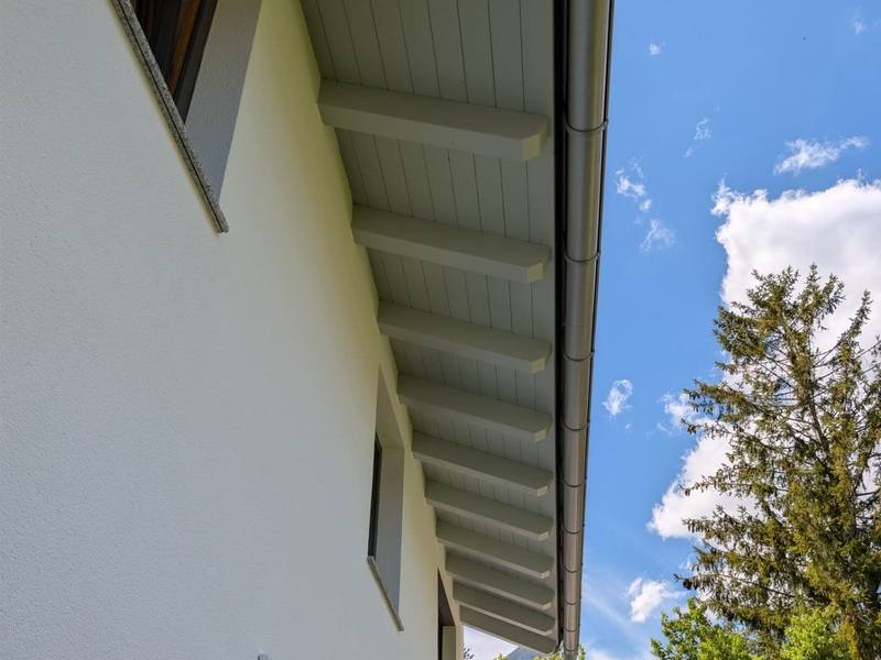 Frisch gestrichene weiße Fassade mit grau lackierter Holz-Dachuntersicht, dahinter blauer Himmel und Tiroler Berge.