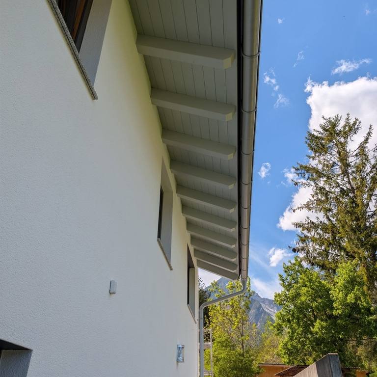 Frisch gestrichene weiße Fassade mit grau lackierter Holz-Dachuntersicht, dahinter blauer Himmel und Tiroler Berge.