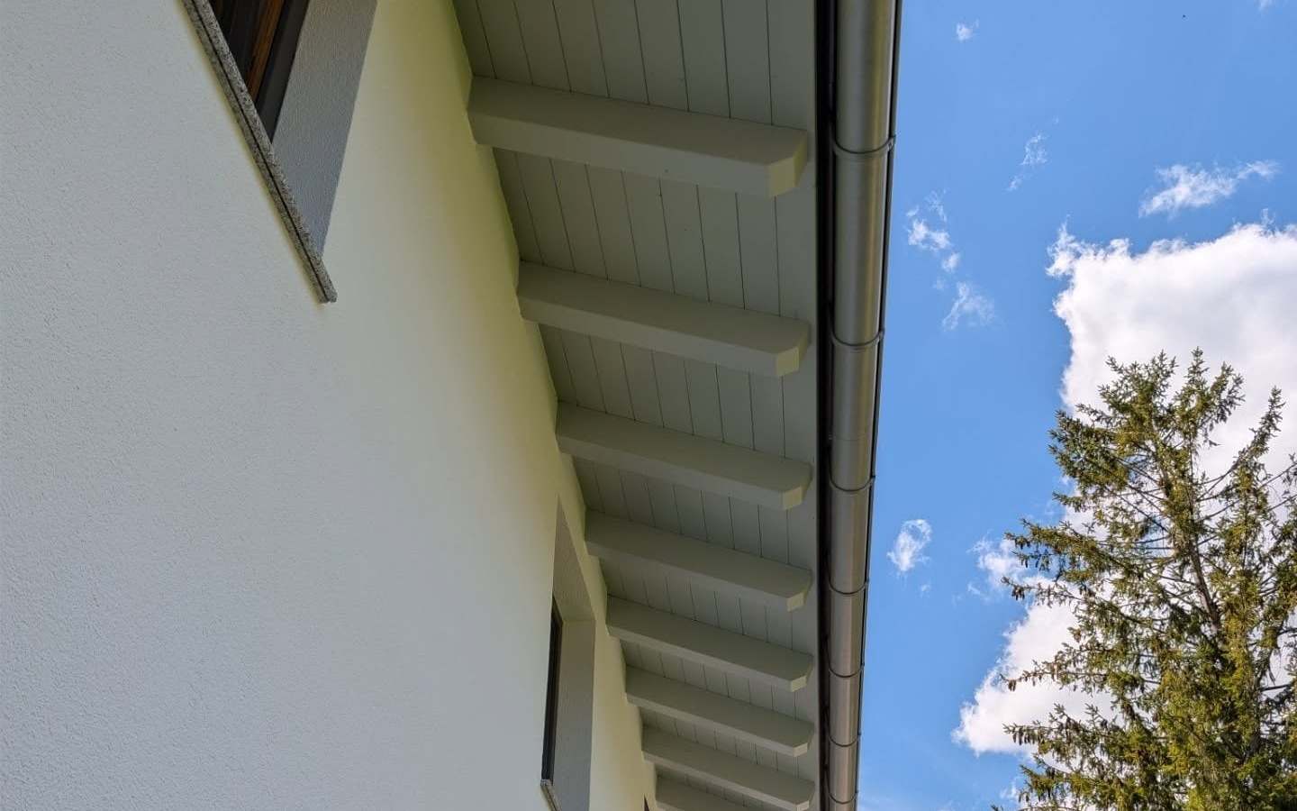 Frisch gestrichene weiße Fassade mit grau lackierter Holz-Dachuntersicht, dahinter blauer Himmel und Tiroler Berge.