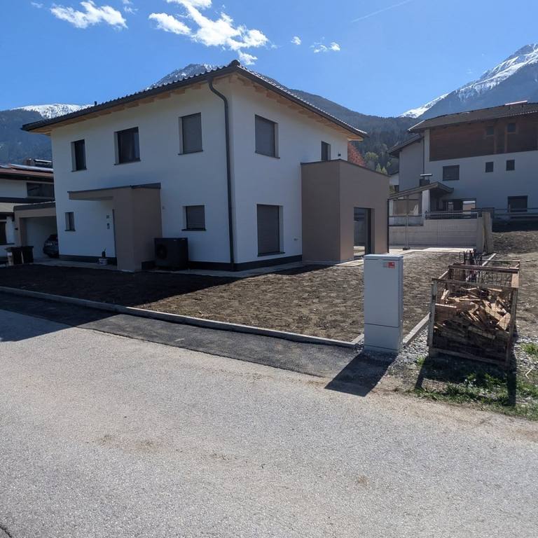 Derselbe Neubau nach dem Anstrich: weiße Fassade mit abgesetztem Farbton, sonniges Wetter, Berge im Hintergrund.