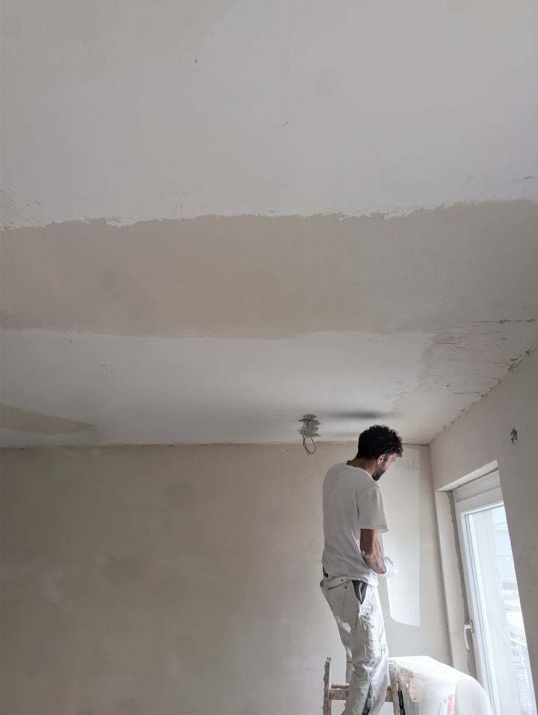 Maler spachtelt im Rohbau von einem Tritt aus die Decke; die frisch abgezogene Fläche zieht sich über den ganzen Raum.