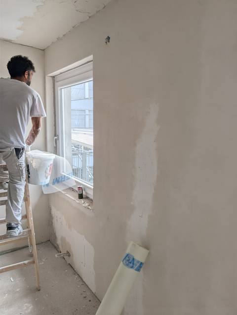 Maler auf einer Leiter spachtelt im Rohbau die Wandfläche neben einem Fenster.