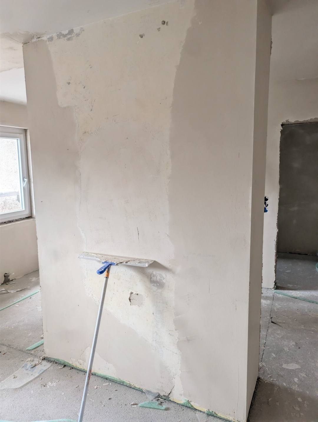 Frisch gespachtelte Wand im Rohbau, davor eine Glättekelle mit Teleskopstiel an der Fläche.