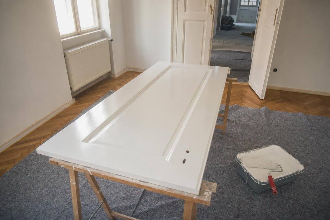 Ausgehängtes Türblatt liegt weiß lackiert auf zwei Böcken in einem abgedeckten Raum, daneben eine Farbwanne mit Roller.