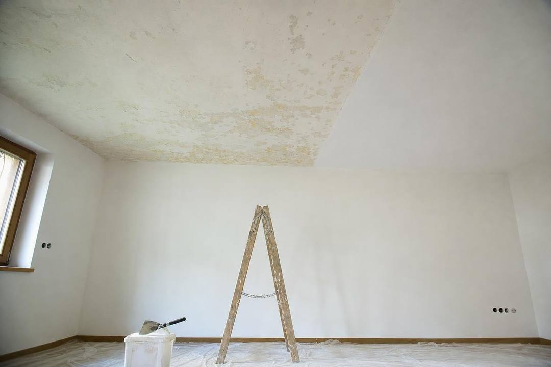 Raumdecke mit großflächigen, gelblichen Feuchtigkeitsrändern und abgeplatzter Farbe, darunter eine Leiter und ein Spachtel.